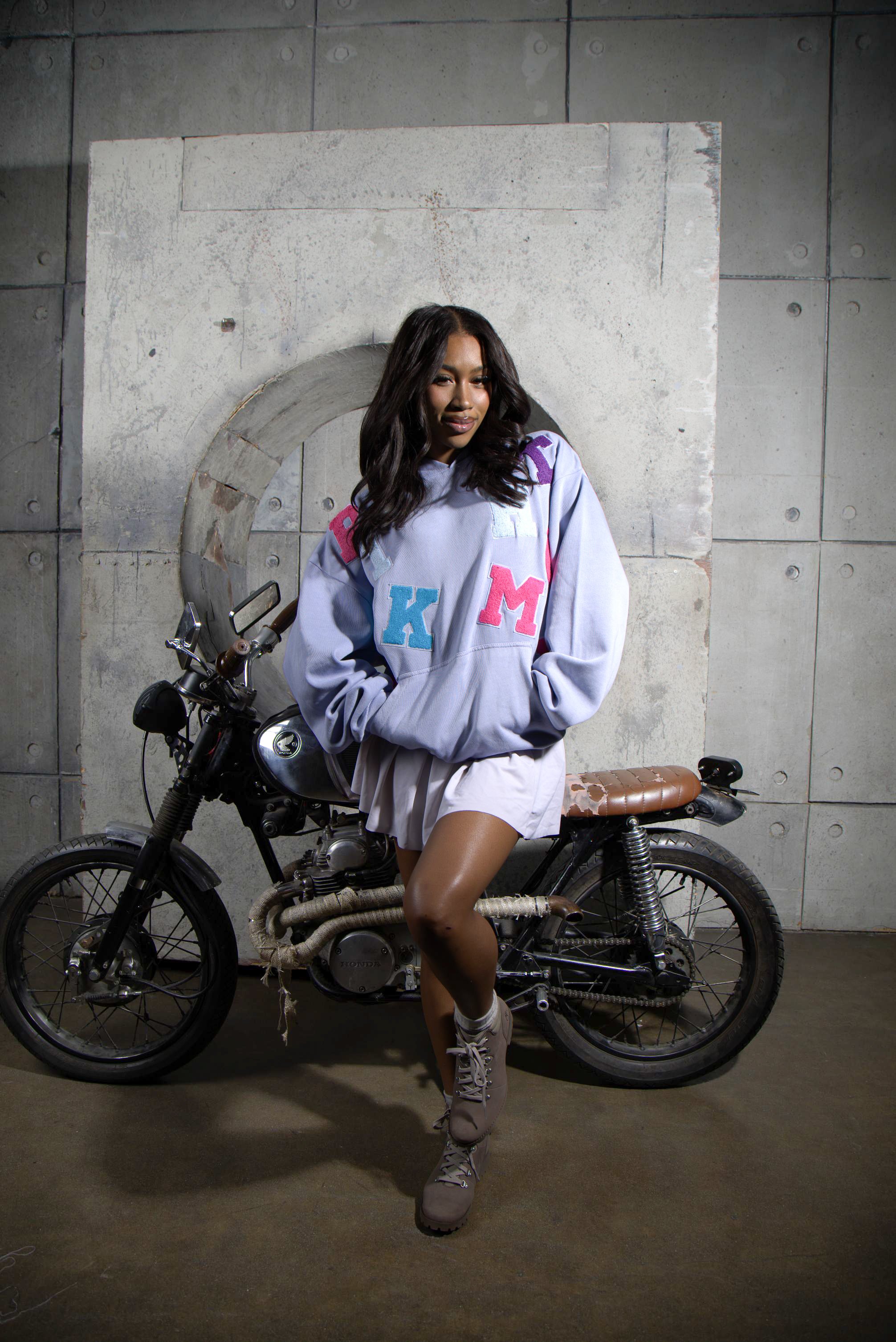 Person wearing a light purple hoodie with colorful letters, standing next to a motorcycle in an urban setting.