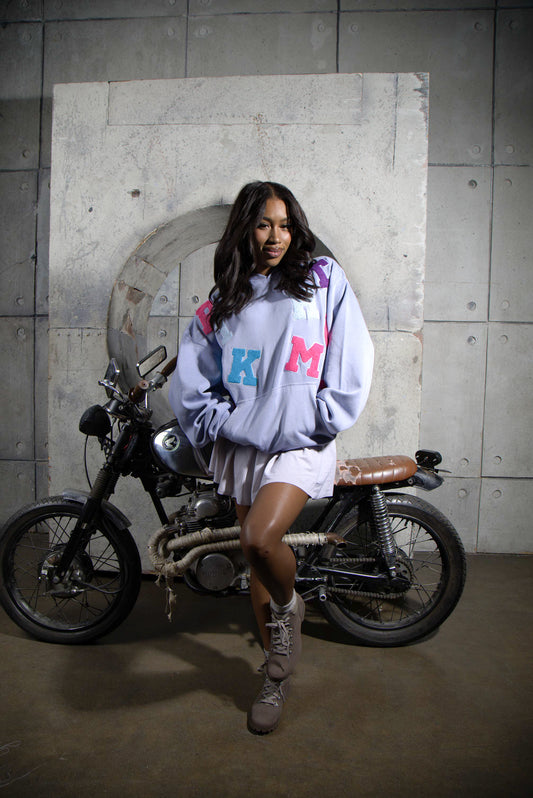 Person wearing a light purple hoodie with colorful letters, standing next to a motorcycle in an urban setting.
